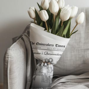 May include: A bouquet of white tulips wrapped in newspaper-style paper. The paper is printed with "The Quincea&ntilde;era Times" and "July 12, 2025 | Miami, FL". The bouquet is tied with a decorative ribbon.