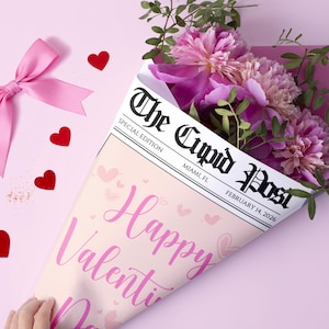 Peut inclure: Un bouquet de pivoines roses envelopp&eacute; dans un journal intitul&eacute; "The Cupid Post" avec le titre "Happy Valentine's Day". Le bouquet est sur un fond rose avec un n&oelig;ud rose et des confettis en forme de c&oelig;ur rouge.