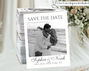 May include: A stack of white "Save the Date" cards featuring a black and white photo of a couple kissing on a beach. The cards include the names "Sophia & Noah" and the date "08.15.2026" with the location "NEW YORK, NY". Text at the top reads "Free Envelopes Included".