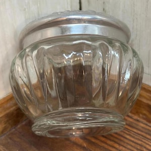 May include: A clear glass jar with a ribbed design and a silver-colored lid. The jar has a rounded shape and sits on a wooden surface. The lid has a decorative pattern around the edge. The jar is empty.