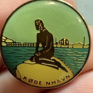 May include: A circular gold-coloured pin depicting the Little Mermaid statue in Copenhagen. The pin has a green and blue backdrop with the text "KØBENHAVN" at the base. Likely a souvenir or collectible item, it is a classic Danish design.