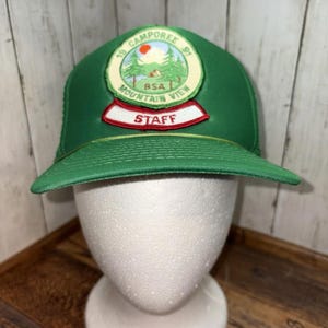 Może przedstawiać: Zielona czapka bejsbolowa z zakrzywionym daszkiem. Czapka posiada naszywkę z napisem "19 Camporee 91 BSA Mountain View STAFF" oraz wizerunkiem góry i drzew. Czapka jest prezentowana na białej głowie manekina.