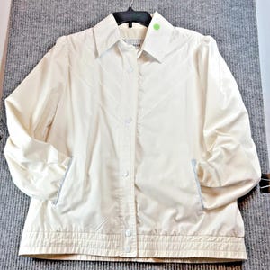 May include: Off-white jacket with a button-up front, collared neckline, and two front pockets. The jacket has a ribbed hem and cuffs, and is hanging on a black hanger. The jacket is made of a lightweight material.