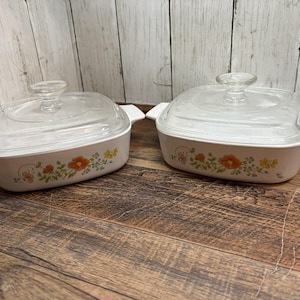 Pode incluir: Duas travessas brancas com tampas de vidro transparente. Cada travessa tem um desenho floral com flores laranja, amarelas e verdes. As travessas retangulares têm alças brancas.