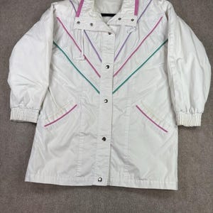 May include: A white windbreaker jacket featuring a high collar, long sleeves with elastic cuffs, and a front snap-button closure. The jacket is accented with thin, V-shaped stripes in pink, purple, and teal across the chest and pockets.