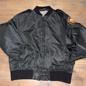 May include: A black bomber jacket featuring a front zipper closure, a ribbed black collar, and a ribbed black waistband. The jacket includes two front pockets with snap buttons and a circular patch with a yellow and red graphic on the left sleeve.