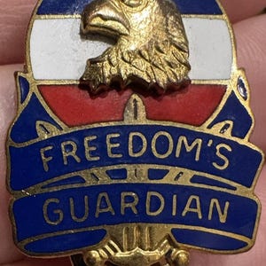 May include: A vintage pin featuring a gold eagle head atop a blue, white, and red design. The pin has the words "FREEDOM'S GUARDIAN" in gold lettering on a blue banner. The pin is round with a gold-colored base.
