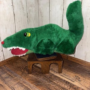 May include: A plush green alligator toy with a red nose, white teeth, and yellow eyes. The toy is sitting on a small wooden stand. The alligator's tail is upright.