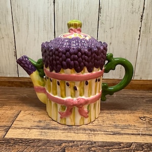 Peut inclure: Une théière décorative en céramique au design fantaisiste. La théière présente un dessus violet texturé, un corps jaune et rose avec des rubans roses et une anse et un bec verts. La théière est posée sur un fond en bois.