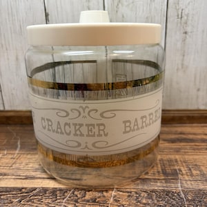 Puede incluir: Un tarro de cristal transparente con tapa de plástico blanca. El tarro presenta una banda horizontal con borde metálico dorado y el texto CRACKER BARREL impreso en gris claro, enmarcado por líneas decorativas de volutas arriba y abajo.