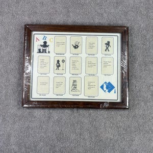 Puede incluir: Un marco rectangular de madera marrón oscuro que contiene un tablero de collage para fotos escolares. El tablero presenta doce secciones etiquetadas desde Kindergarten hasta 12º grado, cada una con espacio para una foto, texto e ilustraciones en silueta.