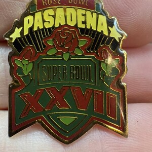 May include: A vintage pin featuring the words "Rose Bowl Pasadena" in yellow and black. The pin also includes the words "Super Bowl XXVII" with red roses and green accents. The pin has a gold border.