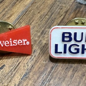 May include: Two vintage Budweiser and Bud Light lapel pins. The Budweiser pin is a red bow tie shape with white text. The Bud Light pin is a white rectangle with blue text and a red border. Both pins have gold-colored pin backs.