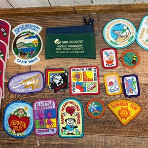 Può includere: Una collezione di toppe Girl Scout e un astuccio verde con cerniera su una superficie di legno. Le toppe presentano vari disegni, tra cui una colomba, una barca a vela e testi come "Summer Fun" e "Health and Fitness."