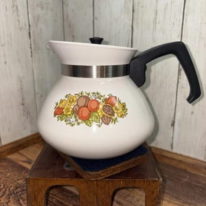 May include: White ceramic teapot with a black handle and lid. The teapot features a decorative band and a floral design with oranges, lemons, and other fruits. The teapot is sitting on a small wooden stand.