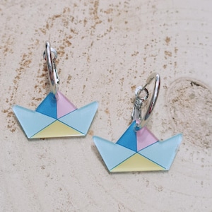 May include: A pair of earrings featuring a paper boat design. Each earring has a geometric pattern in pastel colors: blue, pink, and yellow. The earrings are attached to silver-colored hoop closures. The background is a light-colored, textured surface.