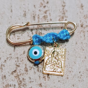 Op de afbeelding: Een gouden veiligheidsspeld met een blauw koord, een blauwe kwade oog-hanger en een gouden hanger die de Maagd Maria en het kindje Jezus voorstelt.