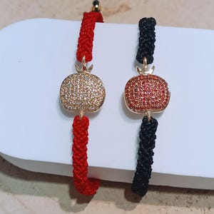 May include: Two adjustable bracelets featuring gold-coloured pomegranate charms. One bracelet has a red woven cord, the other a black woven cord. The pomegranate charms are encrusted with small, sparkling stones.