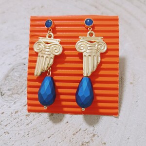 May include: A pair of gold-toned earrings featuring a Greek column capital design at the top, accented with a small round blue stone stud and a larger, faceted, teardrop-shaped blue bead dangling from the base of each column piece.