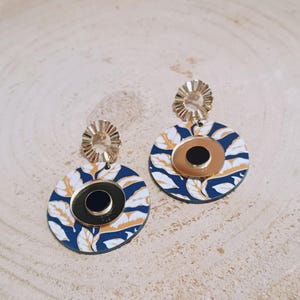 Può includere: Un paio di orecchini color oro con un design circolare. Gli orecchini presentano uno sfondo blu navy con un motivo a foglie bianche e dorate. Ogni orecchino ha una parte superiore color oro e un cerchio nero al centro.