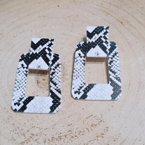 May include: A pair of rectangular earrings with a snake print pattern in black and white. Each earring has a small, clear gem at the top and a rectangular cutout in the center. The earrings are displayed on a light brown wooden surface.