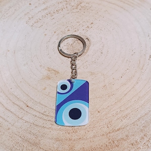 Könnte beinhalten: Ein rechteckiger Schlüsselanhänger mit einem blauen und lila Augendesign. Der Schlüsselanhänger hat einen silbernen Metallring und eine Kette. Das Augendesign zeigt konzentrische Kreise in Blautönen und eine schwarze Pupille. Der Schlüsselanhänger liegt auf einer Holzoberfläche.