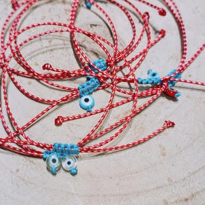Può includere: Diversi braccialetti intrecciati rossi e bianchi con dettagli blu e piccoli ciondoli rotondi blu e bianchi a forma di occhio. I braccialetti sono disposti su una superficie di legno chiaro.