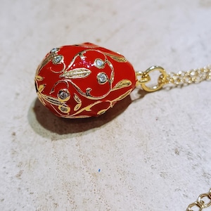 May include: A red egg-shaped pendant with gold filigree and clear crystal accents. The pendant hangs from a delicate gold chain. The chain has a small tag that reads "S-STEEL".