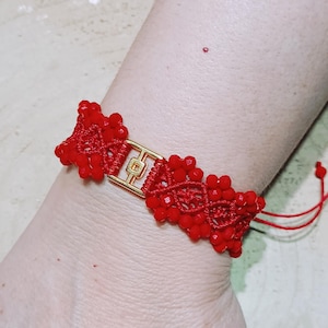 May include: A red woven bracelet featuring small red beads and a central gold-toned rectangular metal charm with a geometric cutout design. The bracelet is constructed with a macrame-style knot pattern and includes adjustable red cord ends.