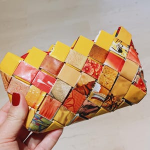 Op de afbeelding: Een geweven clutch-portemonnee gemaakt van gerecyclede materialen. De portemonnee heeft een patchwork-ontwerp met gele, rode en bruine vierkanten. De clutch heeft een rode rits en wordt vastgehouden door een hand met rode nagellak.