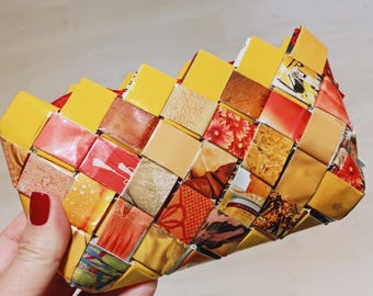 May include: A woven clutch purse made from recycled materials. The purse features a patchwork design with yellow, red, and brown squares. The clutch has a red zipper and is held by a hand with red nail polish.