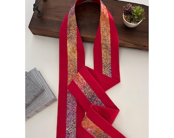 Puede incluir: Una cinta de tela roja con una franja de purpurina dorada y morada en el centro. La cinta está doblada por la mitad y está sobre una superficie de madera.
