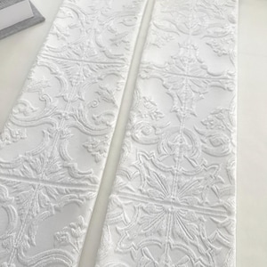 May include: Two white fabric panels with an embossed floral and geometric pattern. The panels are side by side, showcasing the intricate details of the design. The fabric has a textured appearance, with raised floral motifs and geometric shapes.