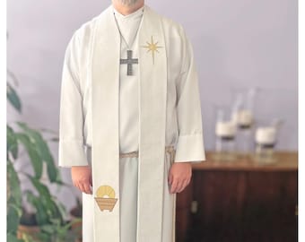 May include: A white liturgical stole with a gold star and a manger scene embroidered on it. The stole is worn by a person with a silver cross necklace.
