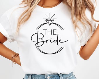 May include: White t-shirt with a black graphic design featuring a diamond ring and the words "THE Bride" in a stylish font. The shirt is made of a soft material and is perfect for a bridal shower or bachelorette party.
