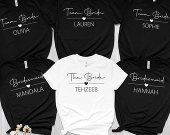 May include: A collection of black and white t-shirts with bridal party text. The shirts feature phrases like "Team Bride" and "Bridesmaid", along with names such as Olivia, Lauren, Sophie, Mandala, and Hannah. One shirt is white and says "The Bride".
