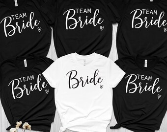 May include: A collection of black and white t-shirts. The shirts feature the words "Team Bride" in a script font with a small heart. The shirts are short-sleeved and suitable for a bridal party.