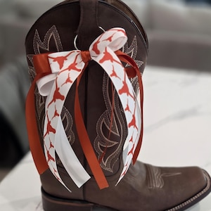 May include: Brown leather cowboy boot with decorative stitching and a square toe. A bow made of white and orange ribbons with a Longhorn pattern is attached to the boot. The boot is a classic Western style.