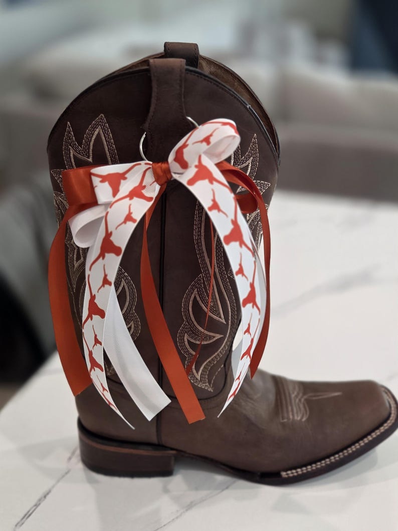May include: Brown leather cowboy boot with decorative stitching and a square toe. A bow made of white and orange ribbons with a Longhorn pattern is attached to the boot. The boot is a classic Western style.