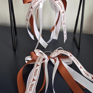May include: Two keychains with bows featuring white and burnt orange ribbons. The ribbons are printed with the word "TEXAS" and a longhorn logo. The bows are attached to a silver metal ring.