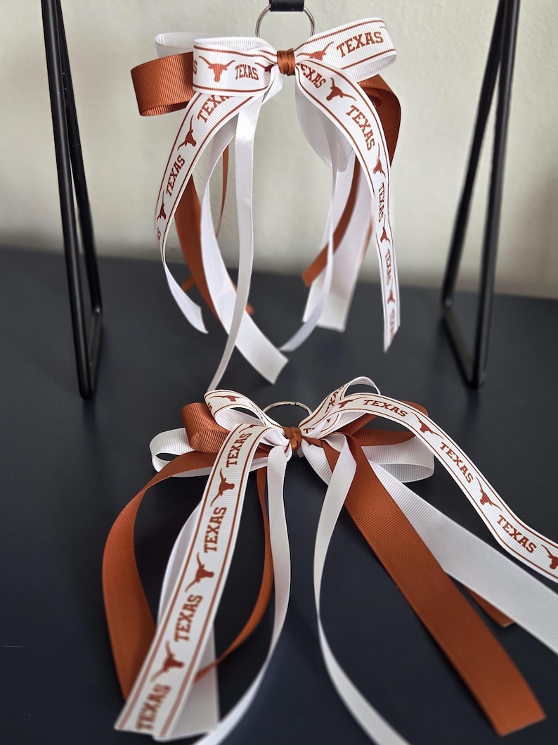 May include: Two keychains with bows featuring white and burnt orange ribbons. The ribbons are printed with the word "TEXAS" and a longhorn logo. The bows are attached to a silver metal ring.