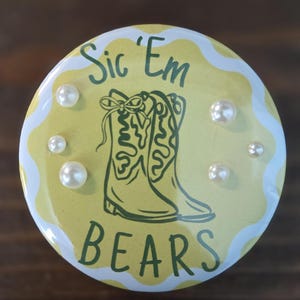 May include: A circular pinback button featuring a pale yellow background with a white wavy border. The center displays a dark green line drawing of a cowboy boot and the text Sic 'Em BEARS. Five small white faux pearls are attached to the surface.