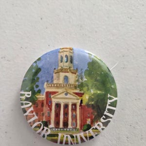 May include: A circular pinback button featuring a watercolor-style illustration of a red brick building with a tall tower and green trees. The text "BAYLOR UNIVERSITY" is printed in white capital letters along the bottom edge of the button.