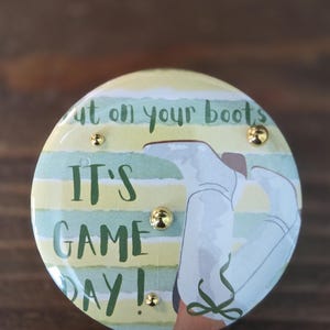May include: A round button featuring a light green and yellow striped background with the text "Put on your boots IT'S GAME DAY!" in dark green. The design includes an illustration of white boots and three small, gold-colored metal studs.