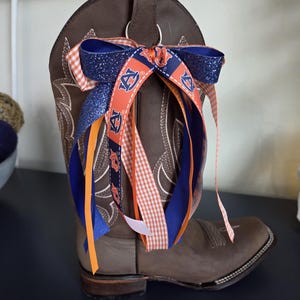 May include: A brown leather cowboy boot with decorative stitching, adorned with a bow made of blue, orange, and white ribbons. The ribbons feature glitter and the letter "A". The boot is displayed on a dark surface.