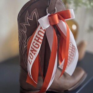 May include: A brown leather cowboy boot with white stitching and a decorative design. Attached is a white and orange ribbon with the word "LONGHORNS" printed on it. The boot has a pull loop and a metal ring.