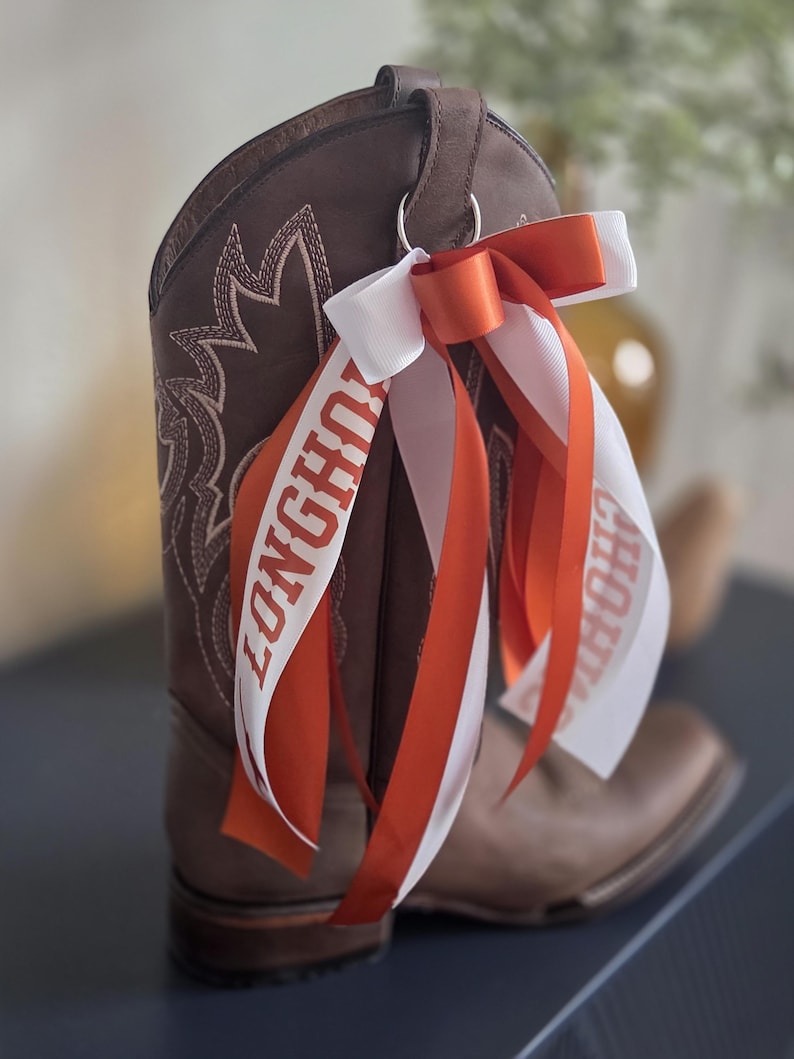 May include: A brown leather cowboy boot with white stitching and a decorative design. Attached is a white and orange ribbon with the word "LONGHORNS" printed on it. The boot has a pull loop and a metal ring.