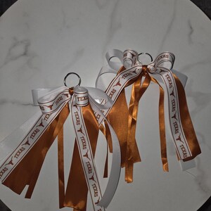 May include: Two keychains with white and burnt orange ribbons. The ribbons are printed with the word "TEXAS" and a Longhorn logo. The keychains are on a white marble surface.