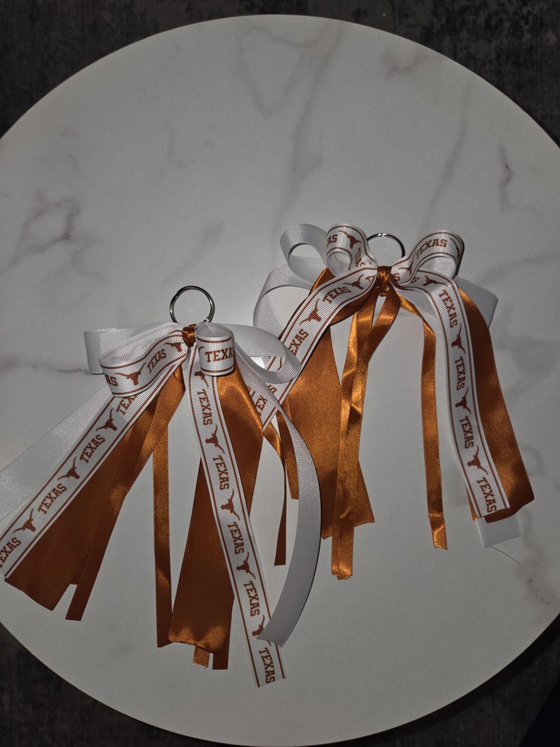 May include: Two keychains with white and burnt orange ribbons. The ribbons are printed with the word "TEXAS" and a Longhorn logo. The keychains are on a white marble surface.