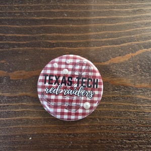 May include: A circular button featuring a red and white gingham pattern background. The text "TEXAS TECH" appears in bold black capital letters above the cursive white text "red raiders". A line drawing of a cowboy boot and three small white pearls.